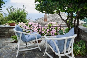 Dos mecedoras blancas con cojines azules, una pared de piedra, arbustos de hortensia rosa y vista al mar.