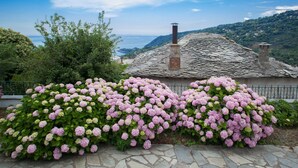 A stone pathway leading to a rustic building with a chimney, surrounded by lush pink hydrangea bushes and a view of the sea.