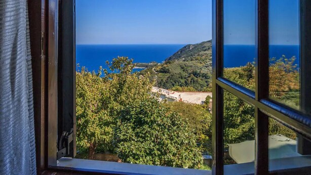 Vista a través de una ventana abierta que muestra un paisaje costero con cielo azul despejado, abundante vegetación y una costa lejana.