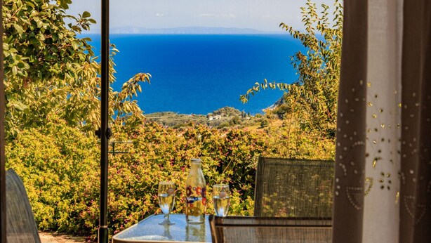A table with two glasses and a bottle, overlooking a scenic view of the sea and hills. - Villa Zina  Studio 1 (Agios Dimitrios Piliou)