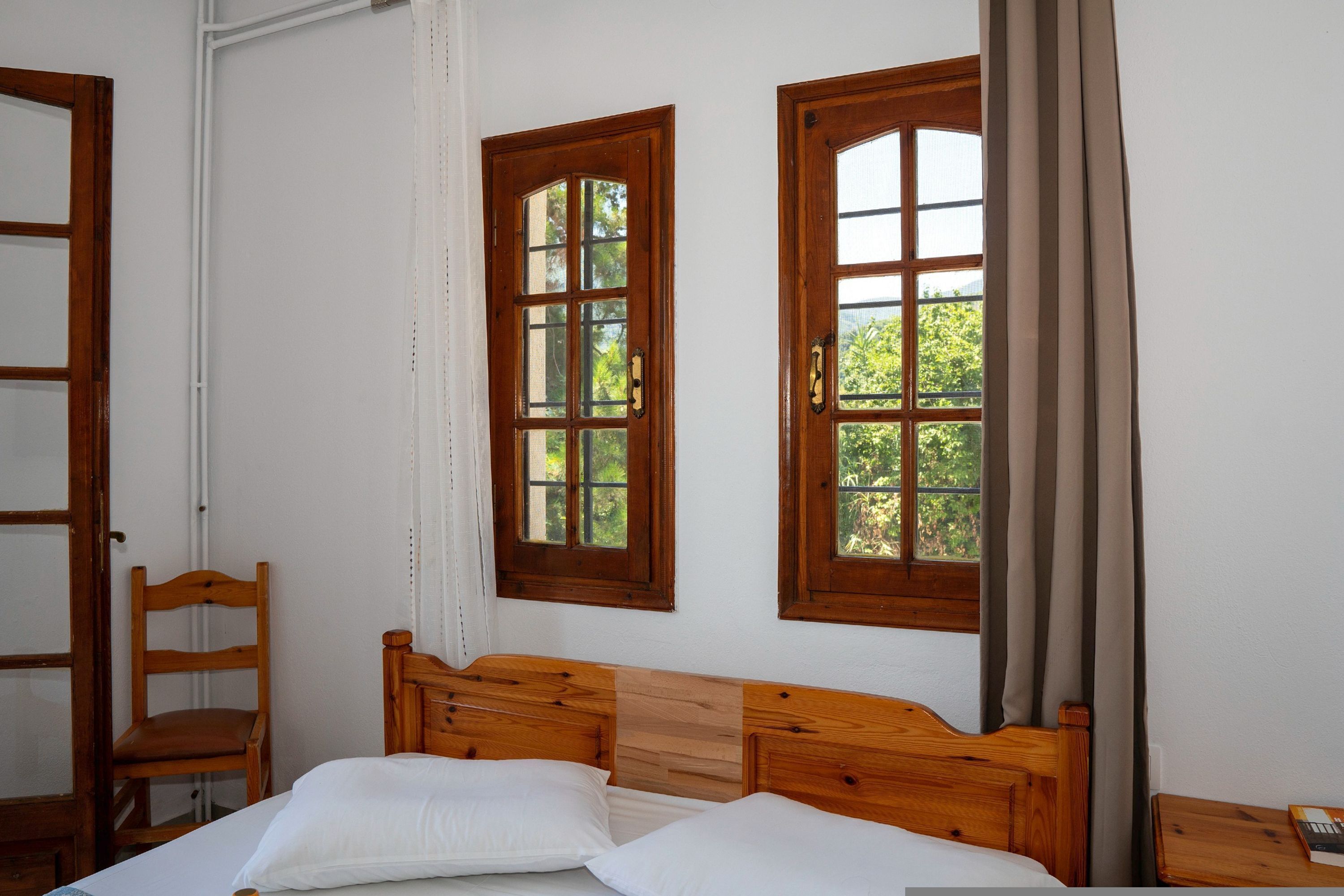 A bedroom with a wooden bed, two windows with curtains, a wooden chair, and a wooden nightstand.
