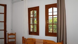 A bedroom with a wooden bed, two windows with curtains, a wooden chair, and a wooden nightstand. - Paradeisos Double Room with Balcony (5) (Agios Dimitrios Piliou)