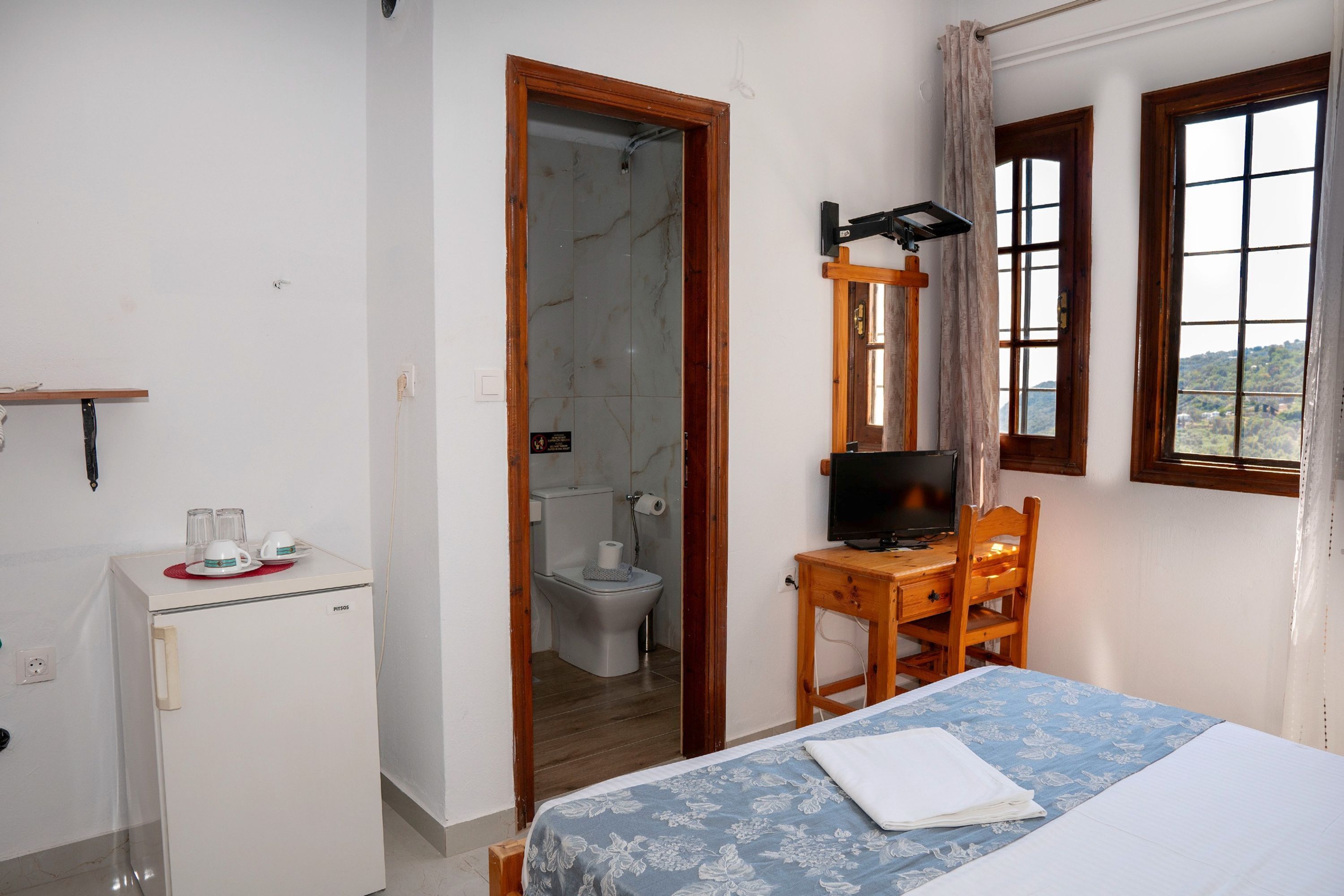 Una camera da letto con un letto, una scrivania con un computer, un piccolo frigorifero e un bagno parzialmente visibile attraverso una porta aperta.