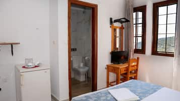 Una camera da letto con un letto, una scrivania con un computer, un piccolo frigorifero e un bagno parzialmente visibile attraverso una porta aperta.