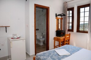 Una camera da letto con un letto, una scrivania con un computer, un piccolo frigorifero e un bagno parzialmente visibile attraverso una porta aperta.