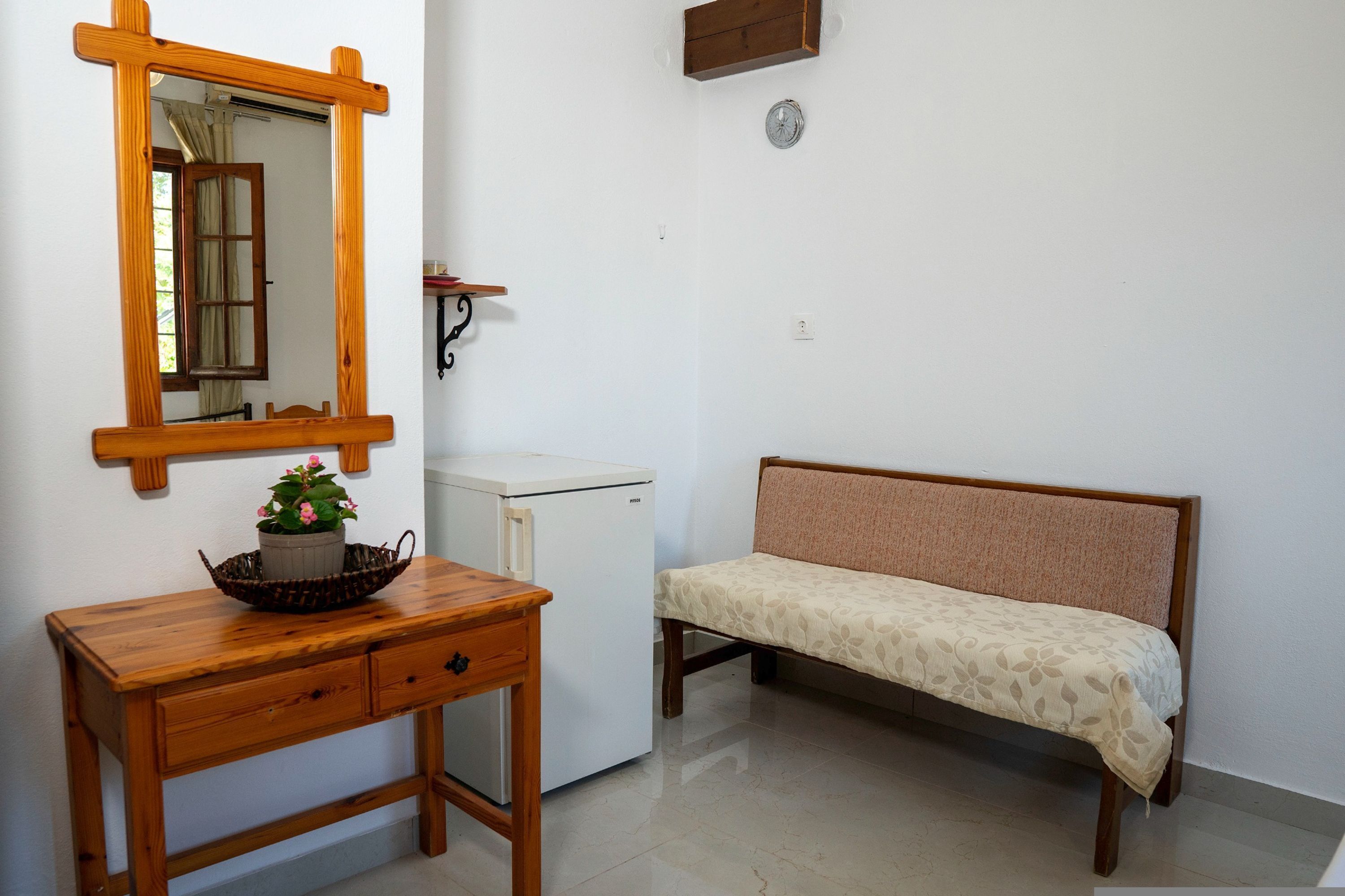 Ein Zimmer mit Schreibtisch aus Holz, einem weißen Kühlschrank, einer Bank, einem Spiegel und einer Wanduhr.