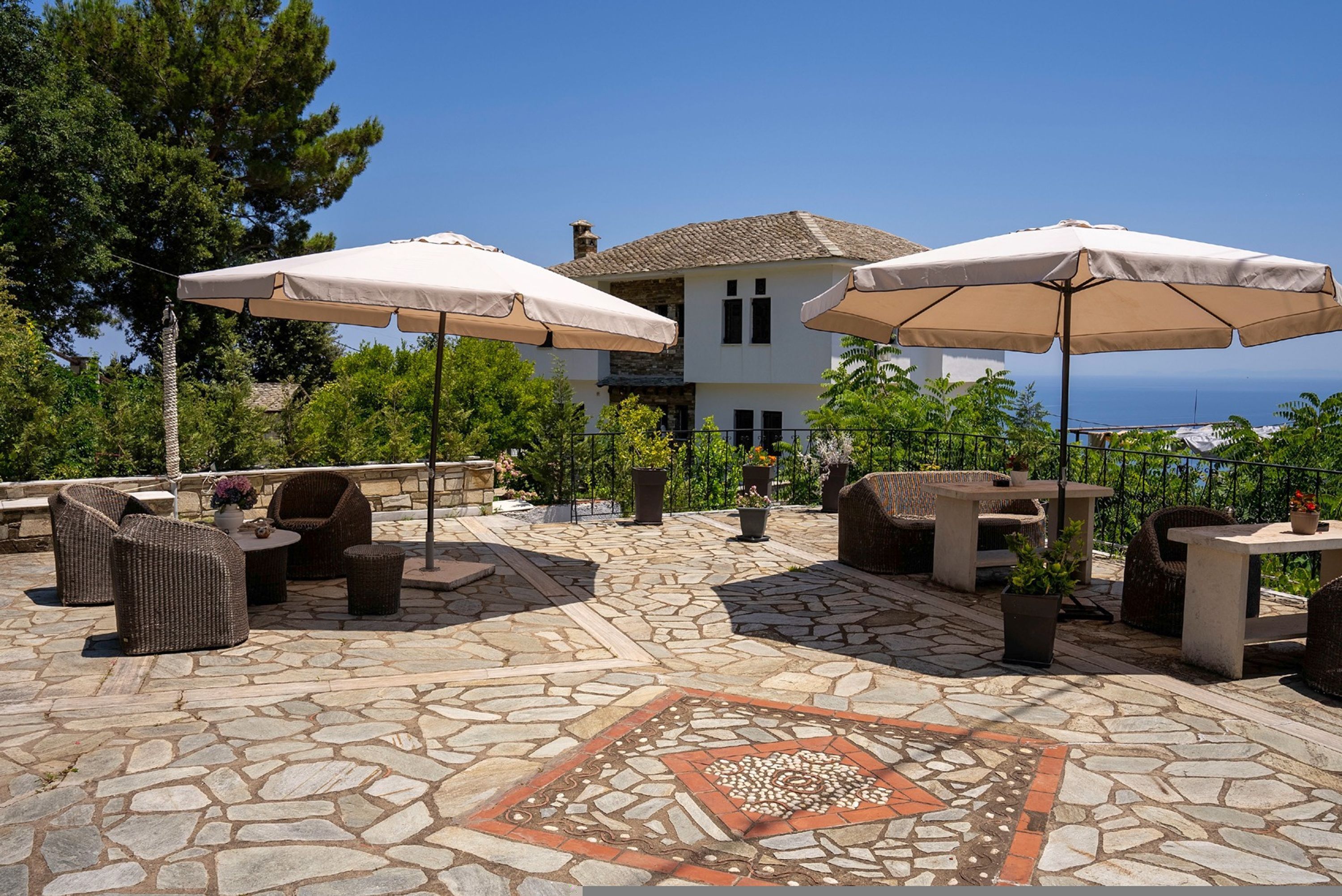 Zona patio con pavimento in pietra, mobili da esterno e ombrelloni, con vista su una casa e del verde.