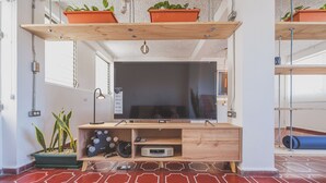 Een moderne woonkamer met een grote flatscreen televisie, houten planken met potplanten, een klein kastje met elektronische apparaten en een rode, patroon vloer.