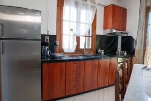 A kitchen with wooden cabinets, a stainless steel refrigerator, a black countertop, and a window with curtains. - Paradeisos Αpartment with Sea View (Agios Dimitrios Piliou)