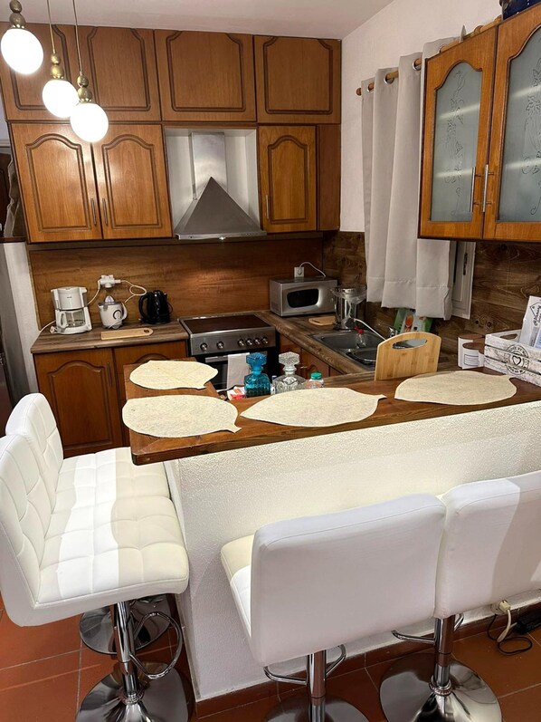 A kitchen with wooden cabinets, a stainless steel range hood, a microwave, and a coffee maker. There are two white bar stools and a wooden table with four placemats. - Harmony Platanidia Pelion (Platanidia)