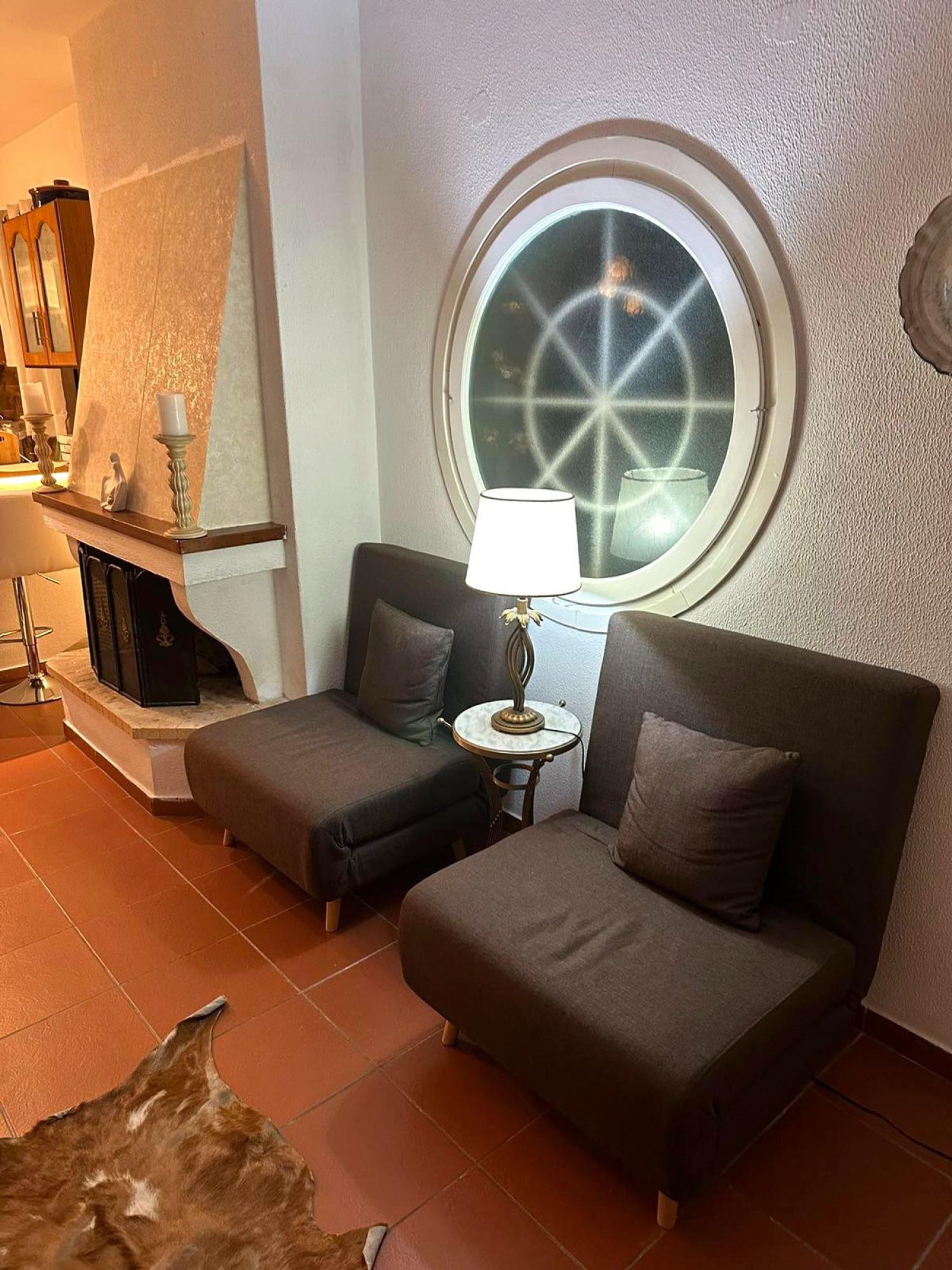 Una habitación acogedora con dos sofás grises, una mesita redonda con una lámpara, una chimenea y un ventanal grande y circular con un diseño de estrellas.