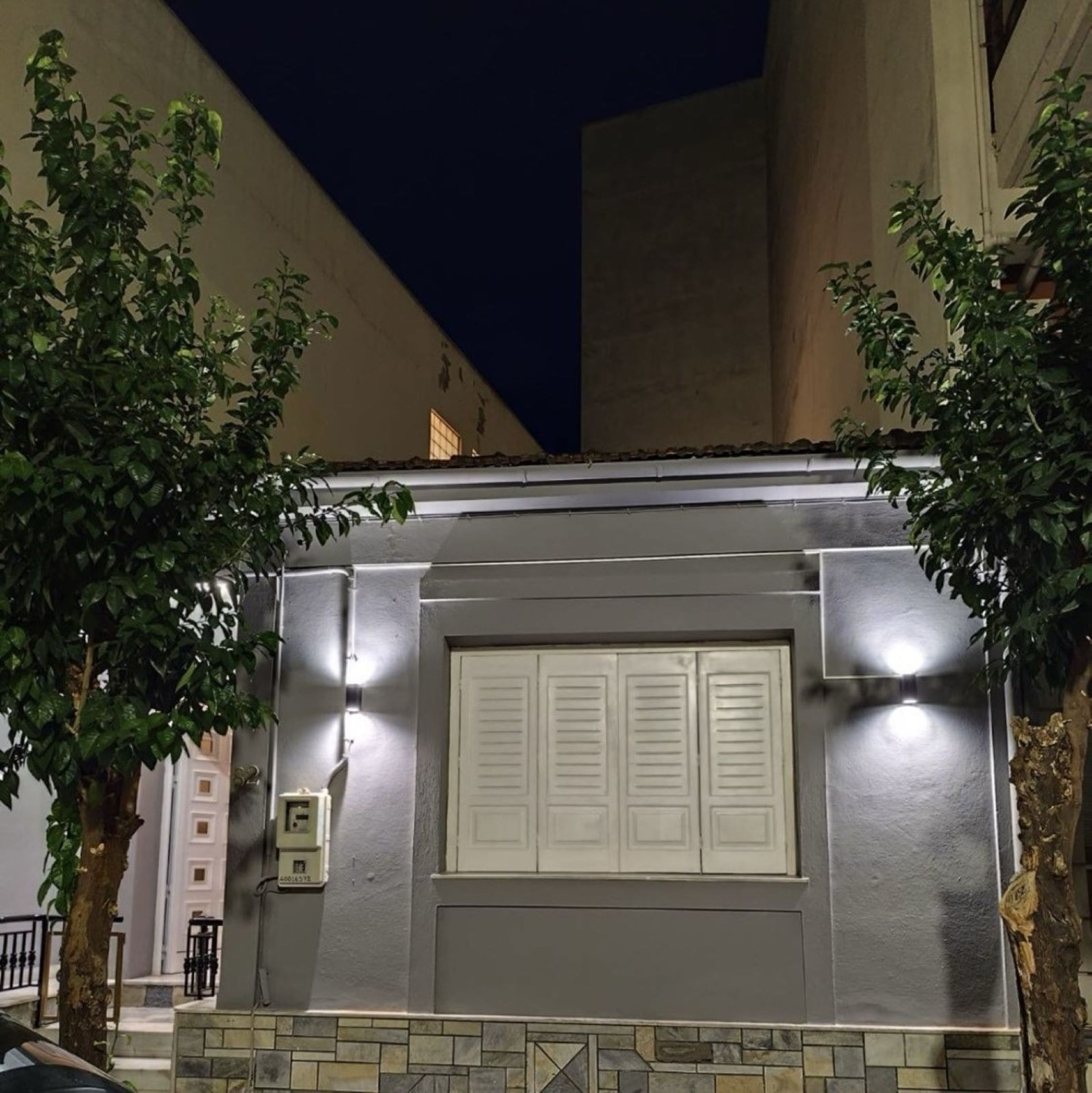 A building with a grey facade, a white door with shutters, and a small window. There are trees in front and a staircase leading up to the entrance.