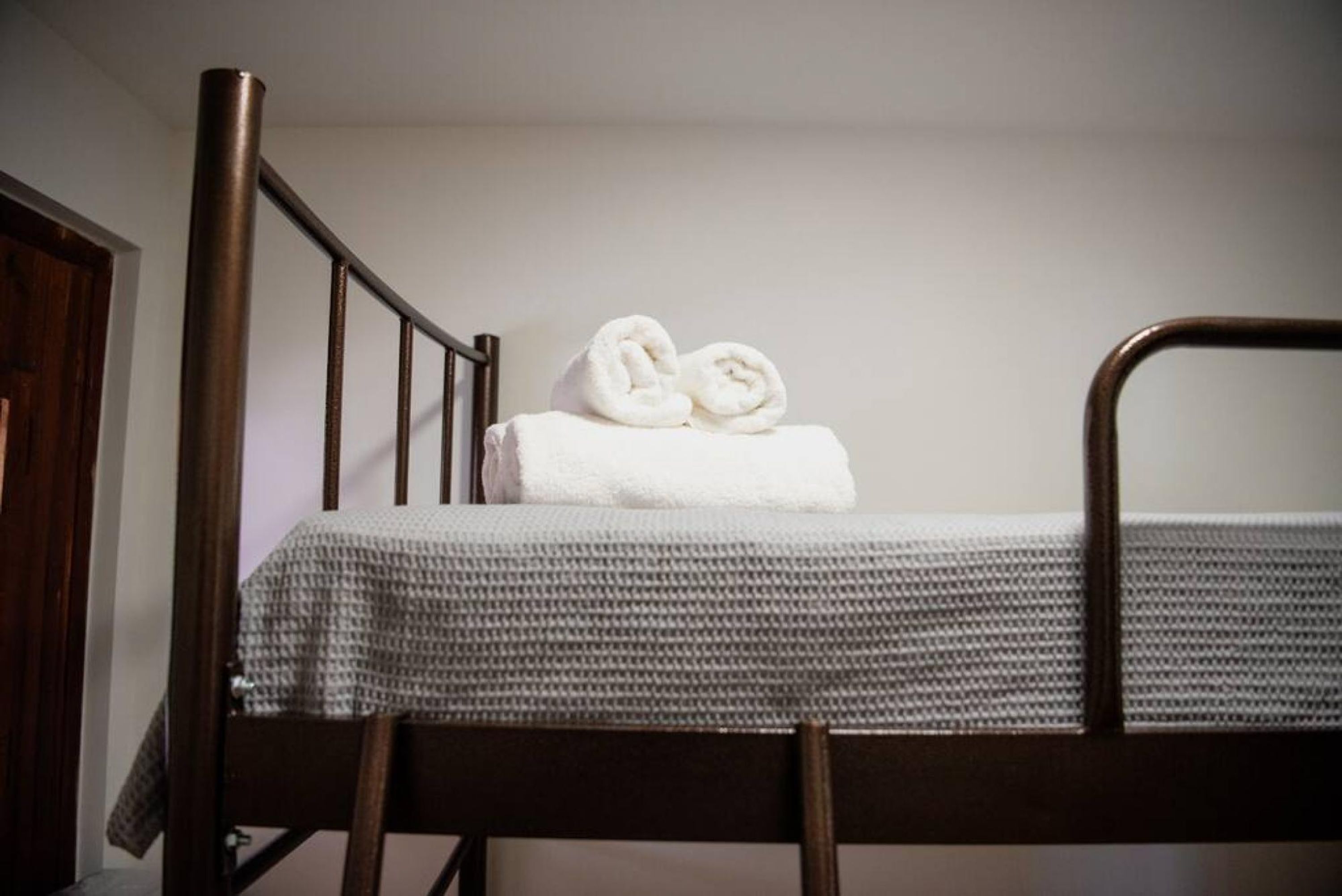 Un lit simple avec un cadre en métal, une couvre-lit à carreaux gris et blancs et deux serviettes blanches roulées posées sur la tête de lit.
