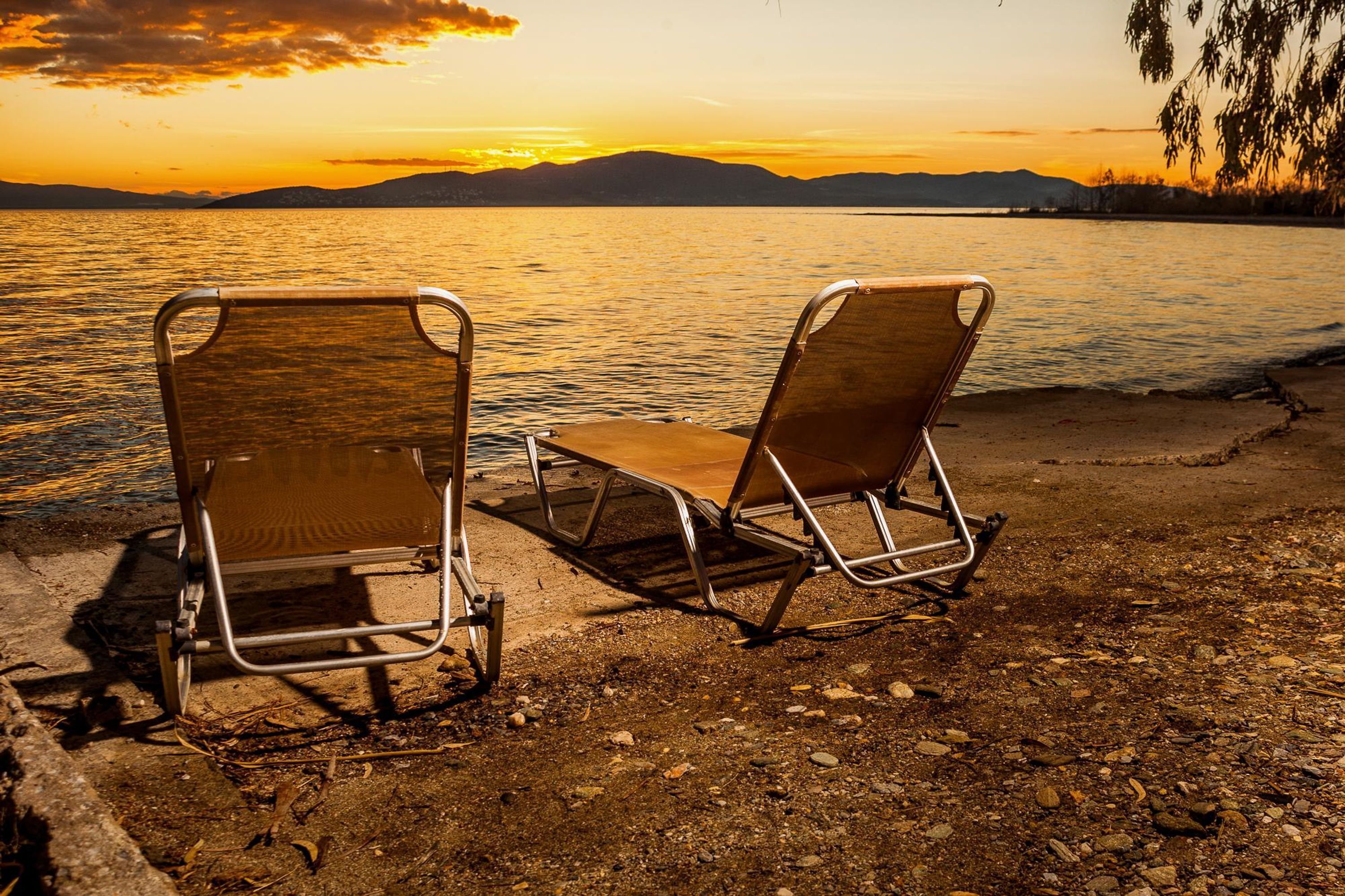 Two metal-framed chairs with wooden seats placed on a rocky shore by a body of water during sunset.