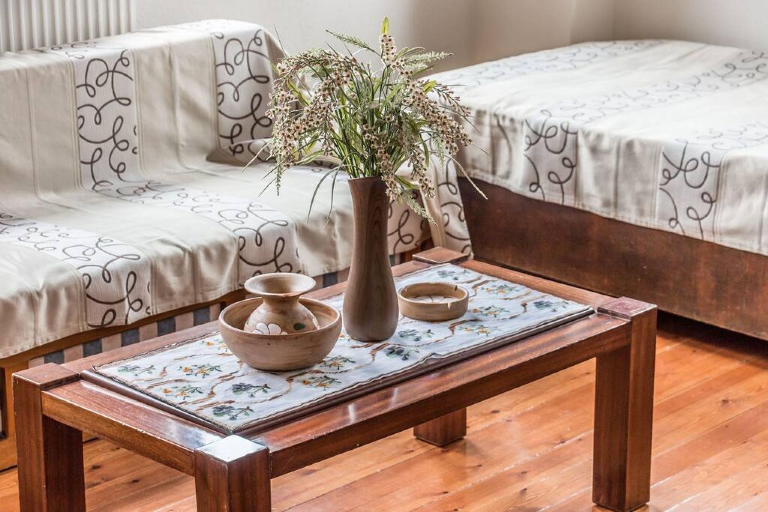 Uma mesa de centro de madeira com um jogo americano floral, um vaso com flores secas e duas tigelas sobre um piso de madeira.