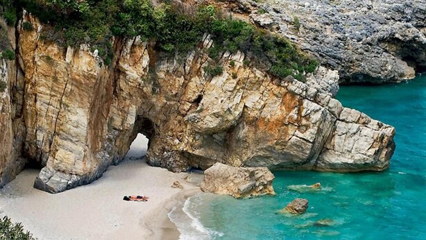 A coastal cliff with a natural arch formation, a sandy beach, and clear turquoise waters.