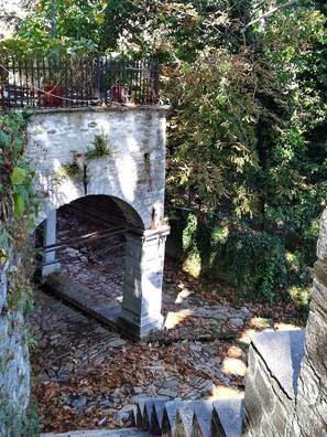 A stone bridge with an arch over a stone pathway, surrounded by dense foliage and a metal fence.