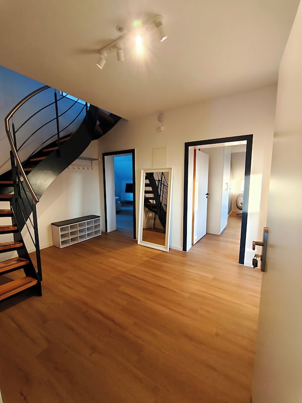 Una habitación amplia con suelo de madera, una escalera en espiral negra y una puerta que da a otra estancia.