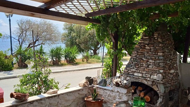 An outdoor patio with a stone-built wood-fired oven, a glass table, wicker chairs, and a view of the sea. - Alonaki Seafront Cottage (Nea Agchialos)