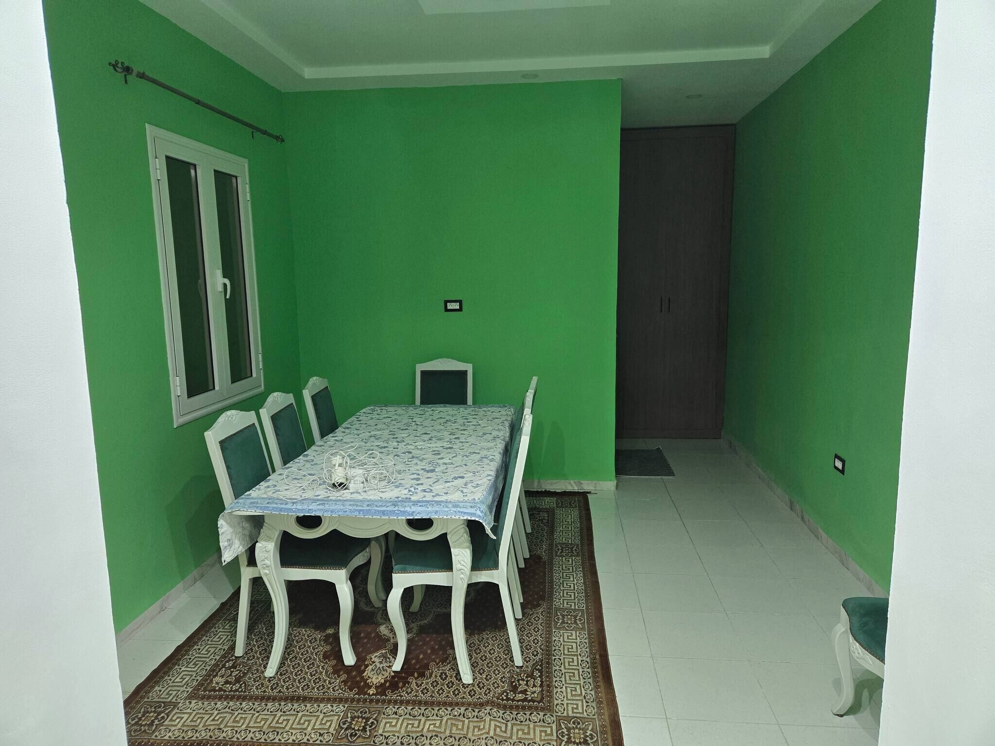 A room with a green wall, a white ceiling, a dining table with chairs, a window, and a door.