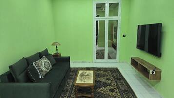 A living room with a green wall, a sofa with patterned cushions, a coffee table, a TV, and a sliding door leading to another room.