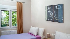 A bedroom with two beds, a window with curtains, a radiator, and a wall-mounted abstract painting. - Aethra House in Agria Volou (Agria Volou)