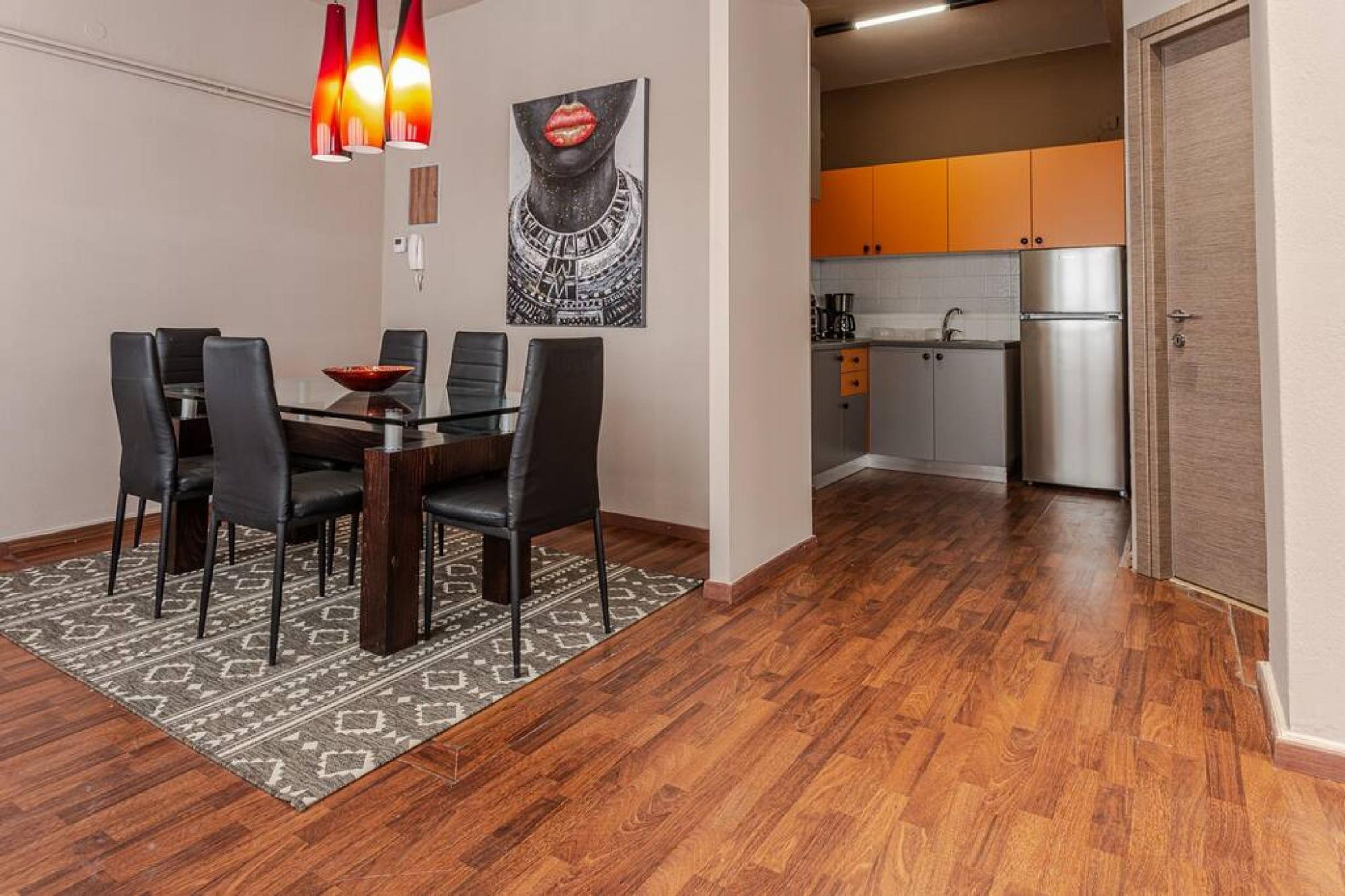 A dining area with a glass table and black chairs, a kitchen with orange cabinets, and a wooden floor.