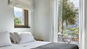 Un dormitorio con cama, almohadas blancas, ventana con vistas a zonas verdes y montañas, y balcón con mesa y sillas.