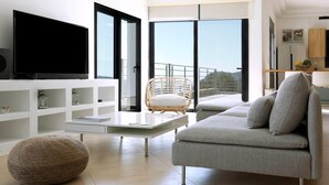 A modern living room with a sofa, a television, a wicker ottoman, and a view of the outdoors through large windows. - Villa Aires with Private Pool (Vrachasi)