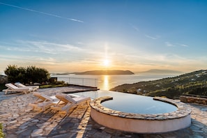 A pool with lounge chairs, a stone deck, and a view of the sea and hills.
