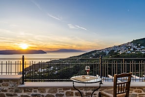 A balcony with a table, a chair, a glass of red wine, and a view of a coastal town at sunset.