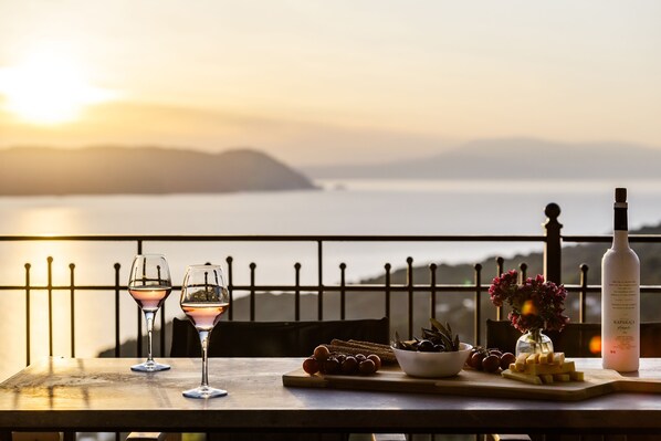 A table with two glasses of wine, a bottle of wine, a bowl of olives, and a vase of flowers, overlooking a body of water and mountains.