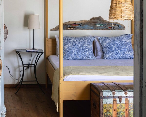 A bed with a wooden frame, blue patterned pillows, a woven headboard, a bedside table with a lamp, and a rustic trunk. - Tinos experience: the house (Panormos)