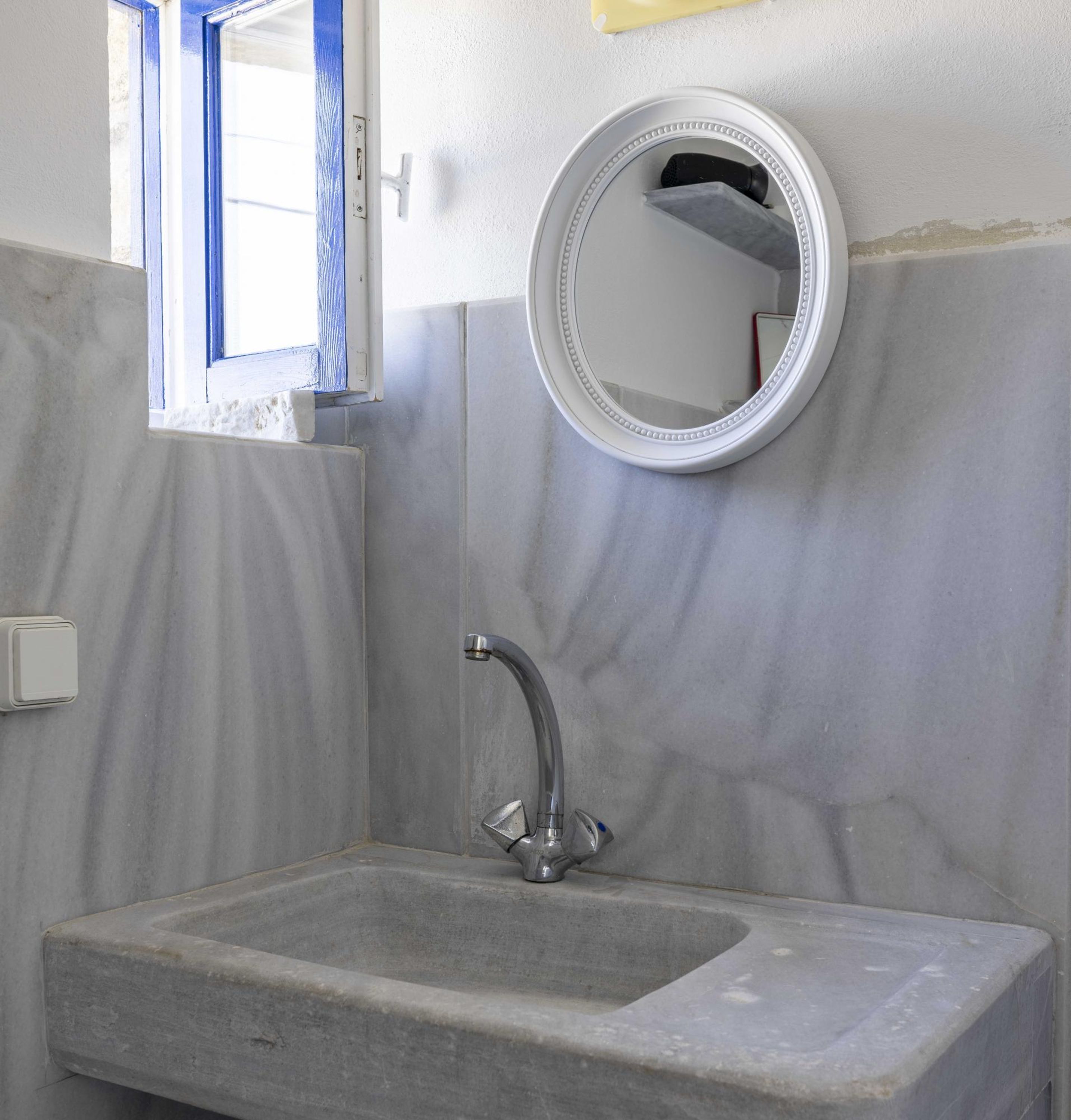 Een badkamer met een marmeren wastafel, een ronde spiegel en een raam met blauwe omlijsting.