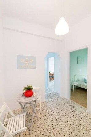 A room with a white wall, a small round table with a red pot, a white chair, and a hanging light fixture. - Mimosa: The mosaic home (Tinos)