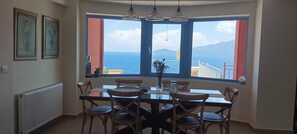 Ein Essbereich mit Tisch und Stühlen, einer Blumenvase und Blick auf das Meer durch große Fenster.