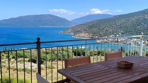 Un balcon avec une table et des chaises, donnant sur un paysage côtier avec des montagnes et une étendue d’eau.