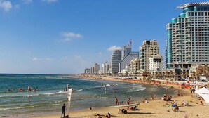 A beach with people swimming and sunbathing, a pier, and tall buildings in the background. - Hayarkon Blue Sea I in Tel Aviv-Yafo (Tel Aviv-Yafo)