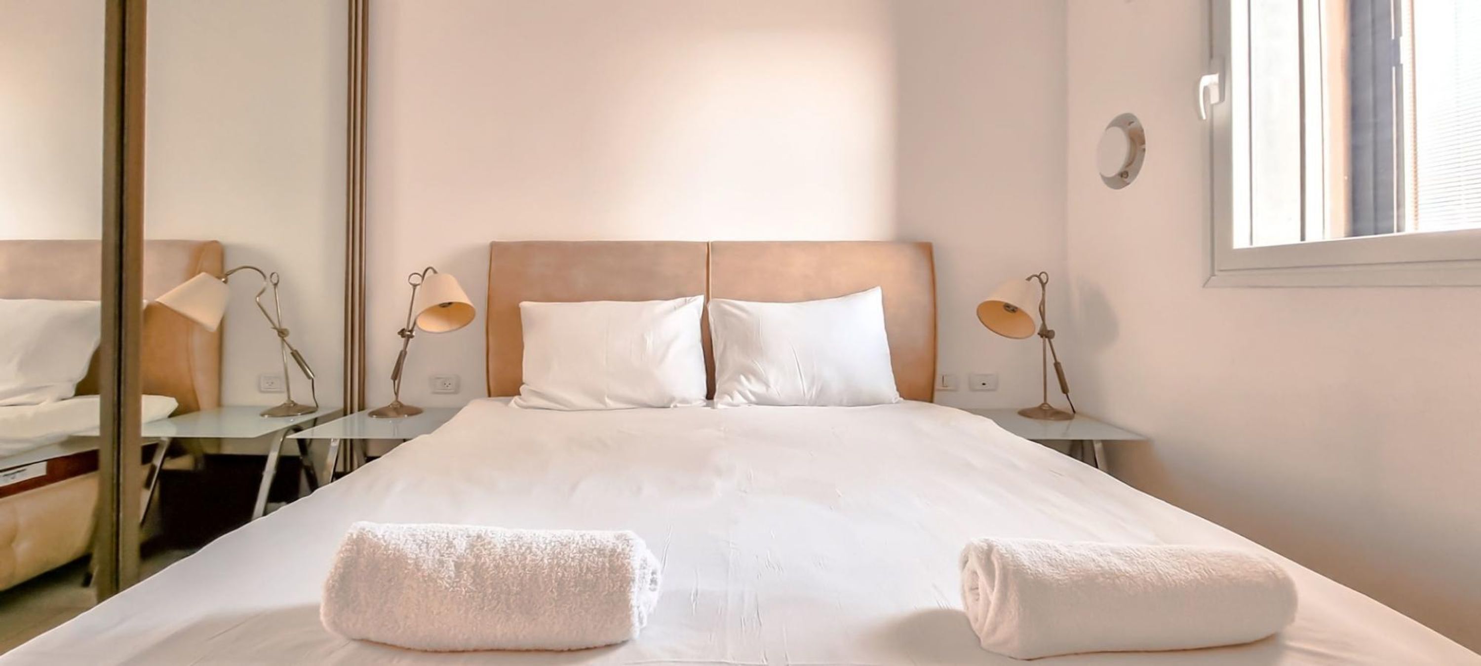 Un lit bien fait, avec du linge de lit blanc et deux serviettes, une table de chevet avec une lampe, et un grand miroir au mur.