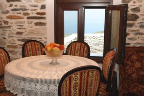 A round table with a crocheted tablecloth, surrounded by six patterned chairs, with a bowl of fruit on the table and a stone wall in the background.