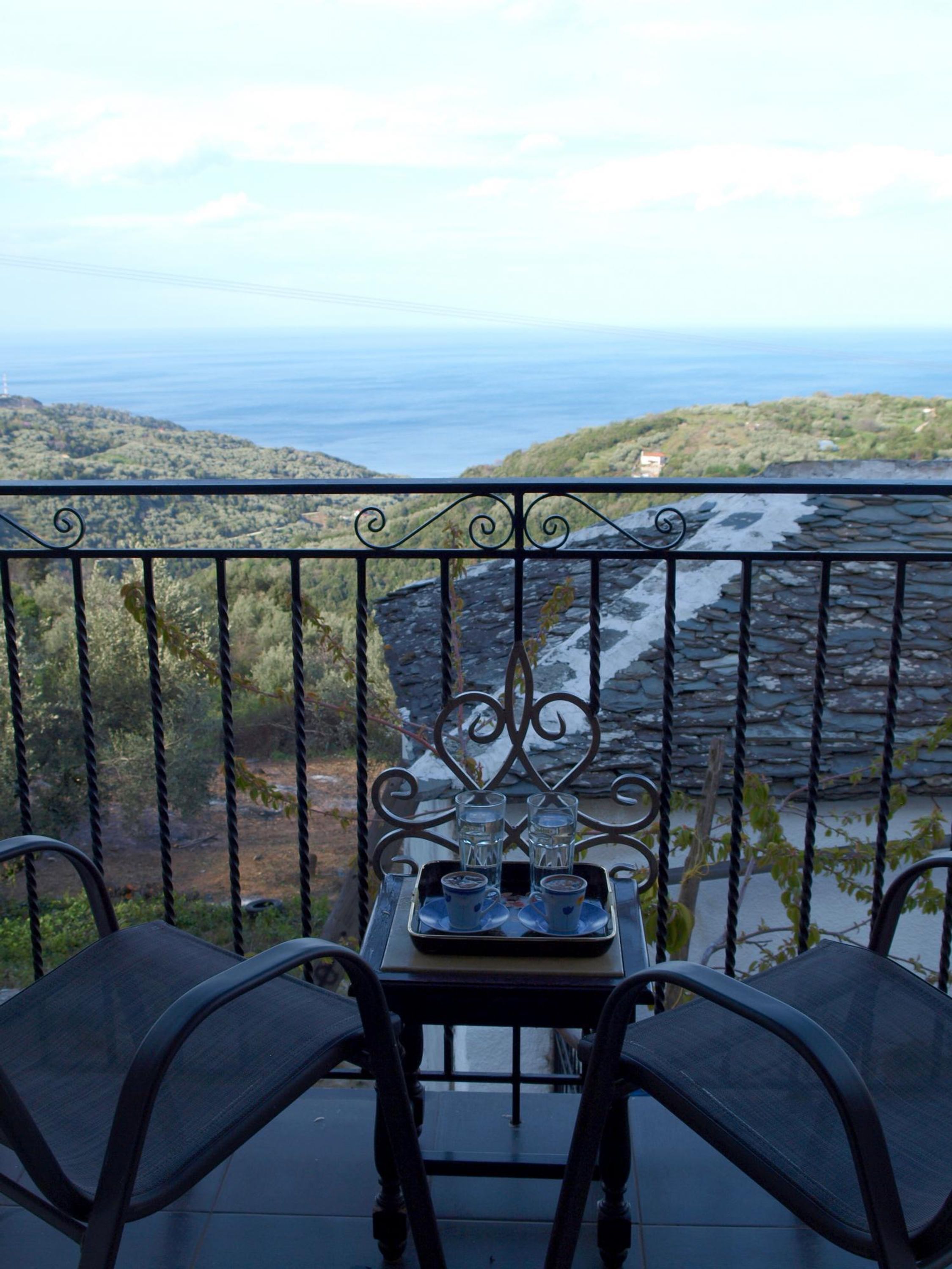 Un balcone con tavolo e sedie, affaccia su un paesaggio costiero con colline e mare.
