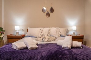 Siististi pedattu sänky, jossa on violetti peitto, kaksi yöpöytää, joissa on lamput, sekä koristeelliset seinävaatteet.