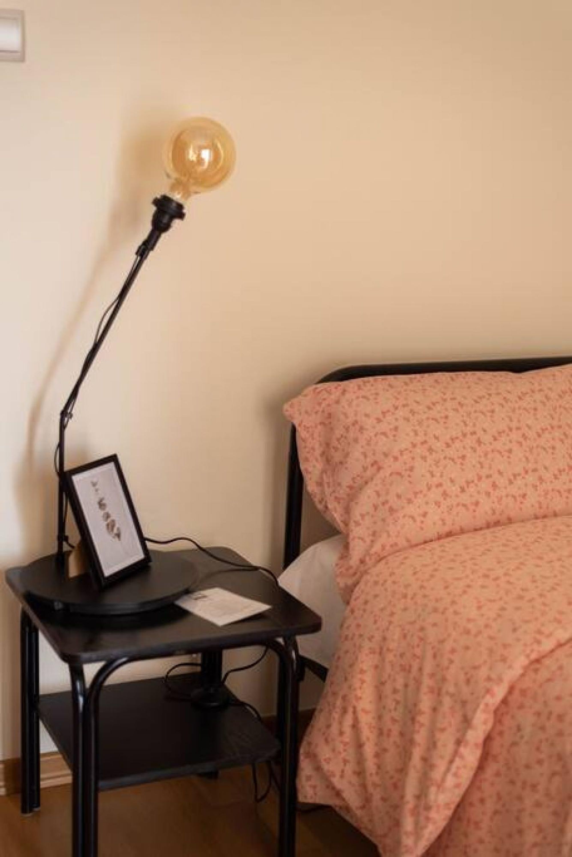A bed with a floral bedspread, a bedside table with a lamp, and a framed picture on the table.