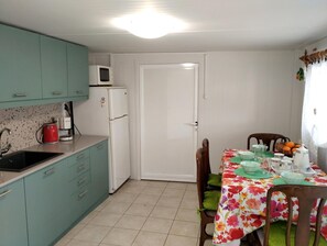 Eine kompakte Küche mit türkis-blauen Schränken, einem weißen Kühlschrank, einer Mikrowelle und einer Kaffeemaschine. Ein Essbereich mit einer blumigen Tischdecke und Stühlen ist ebenfalls zu sehen.