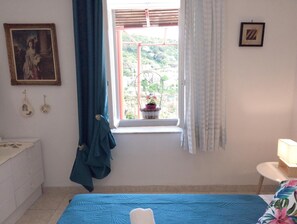 Ein Schlafzimmer mit Bett, einem Fenster mit Vorhängen, einem Schminktisch, einem Gemälde und einem kleinen Tisch mit Lampe.