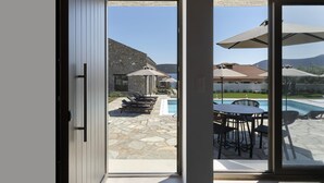 A view through a glass door to a pool area with lounge chairs, umbrellas, and a stone patio.