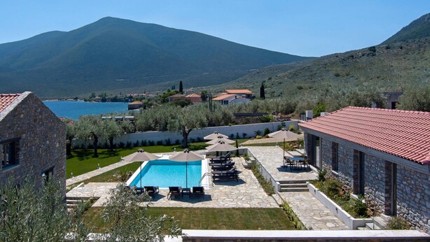 A pool area with lounge chairs and umbrellas, surrounded by stone buildings and a view of a lake and mountains.