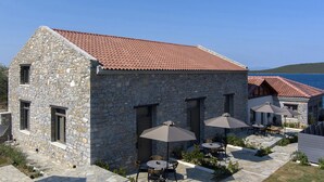 Un edificio de piedra con techo de tejas rojas, mesas al aire libre con sombrillas y vista al mar.