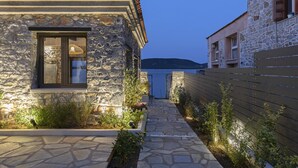A stone building with a tiled roof, a window with a dark frame, and a pathway leading to a gate. - Lichnos Luxury Suite (Sourpi)