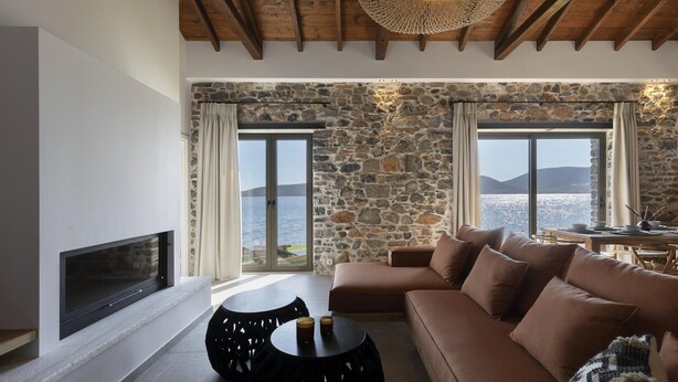 A living room with a stone wall, a sofa, a fireplace, and a view of the sea. - Lichnos Luxury Suite (Sourpi)
